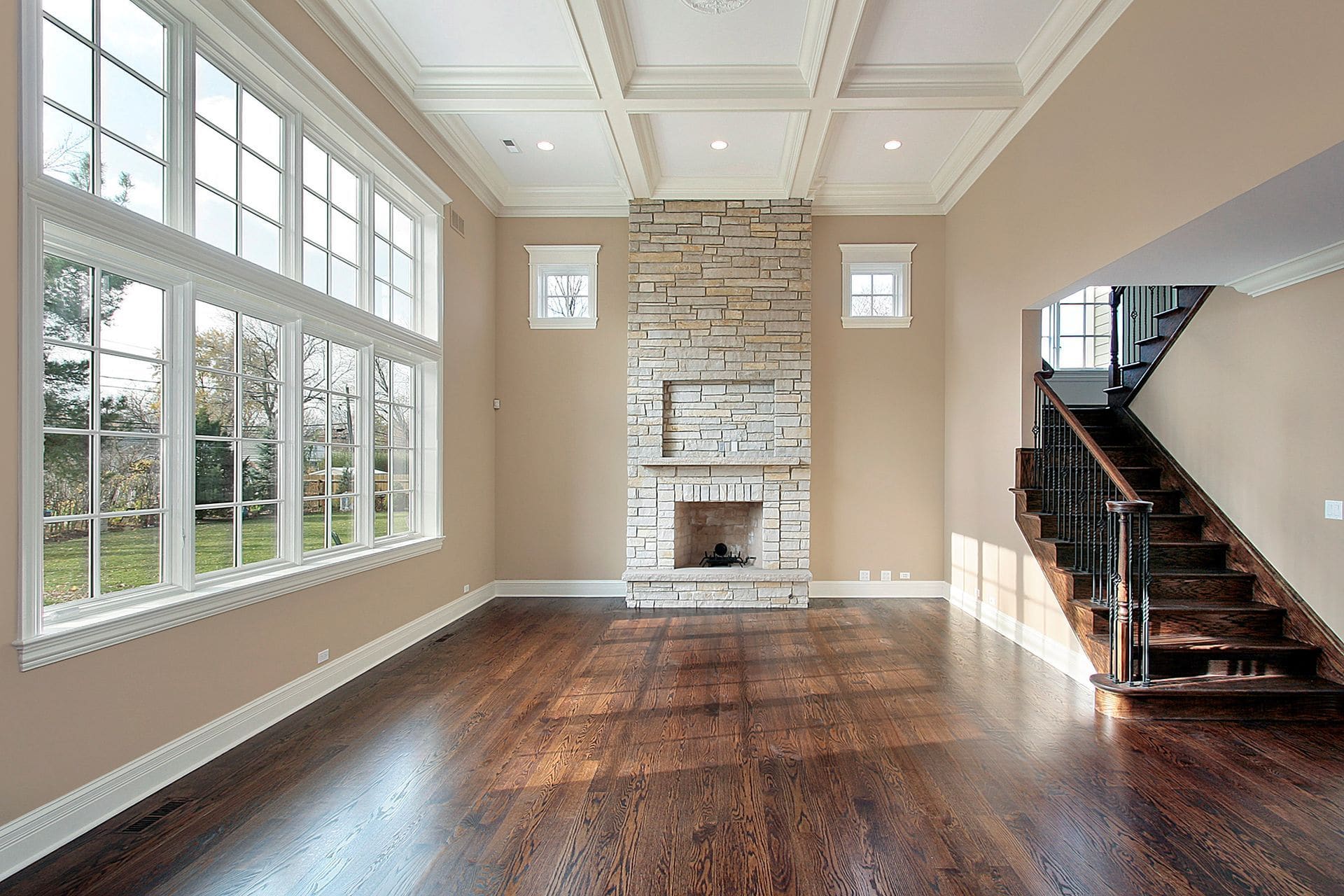 Spacious living room with fireplace, hardwood floors, coffered ceiling, large windows, and wooden staircase