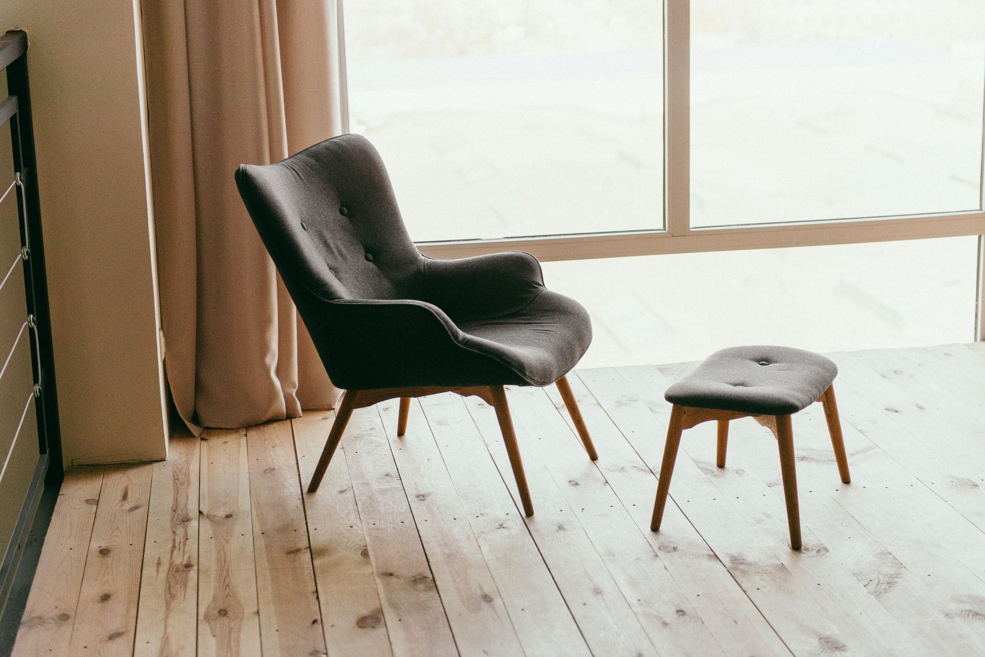 Modern dark gray armchair with wooden legs and matching ottoman near large window with beige curtains